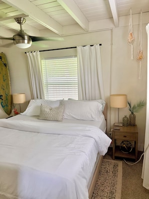 Un dormitorio con cama, dos mesitas de noche, ventilador de techo, ventana con cortinas y una tabla de surf en la pared.