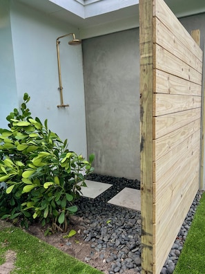 A modern outdoor shower area with a wooden privacy screen, a concrete wall, and a small garden with green foliage and pebbles. - The Surf Shack - Minutes from Beach and Atlantic Ave (Boynton Beach)