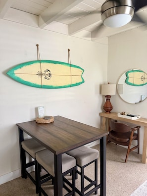 Una habitación con una mesa de madera, dos sillas, un camino de mesa tejido, una decoración de tabla de surf, una lámpara, un espejo y un pequeño estante con objetos.