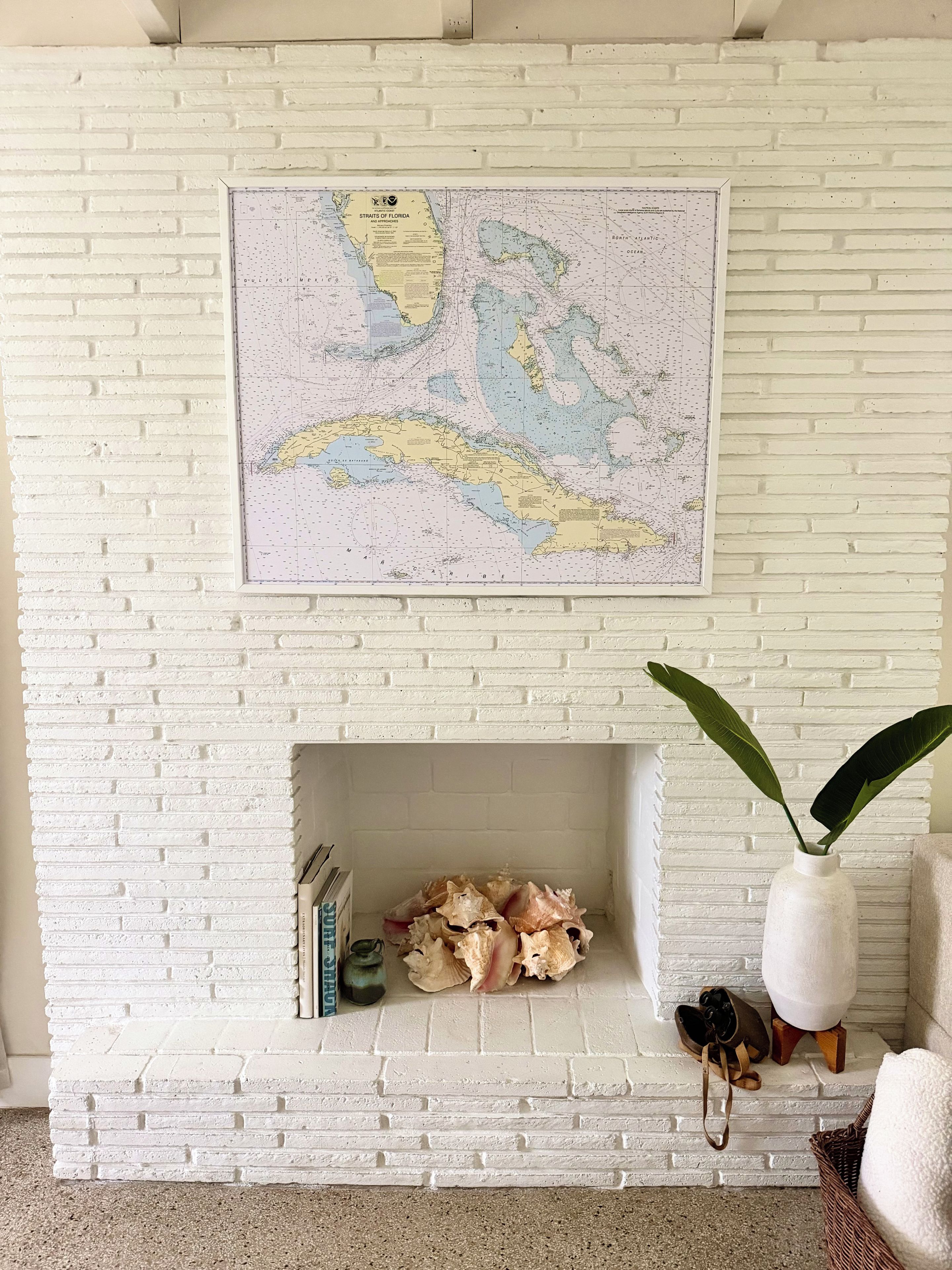 A white brick fireplace with a map above it, a plant in a white vase, and a pair of shoes on a small stool.