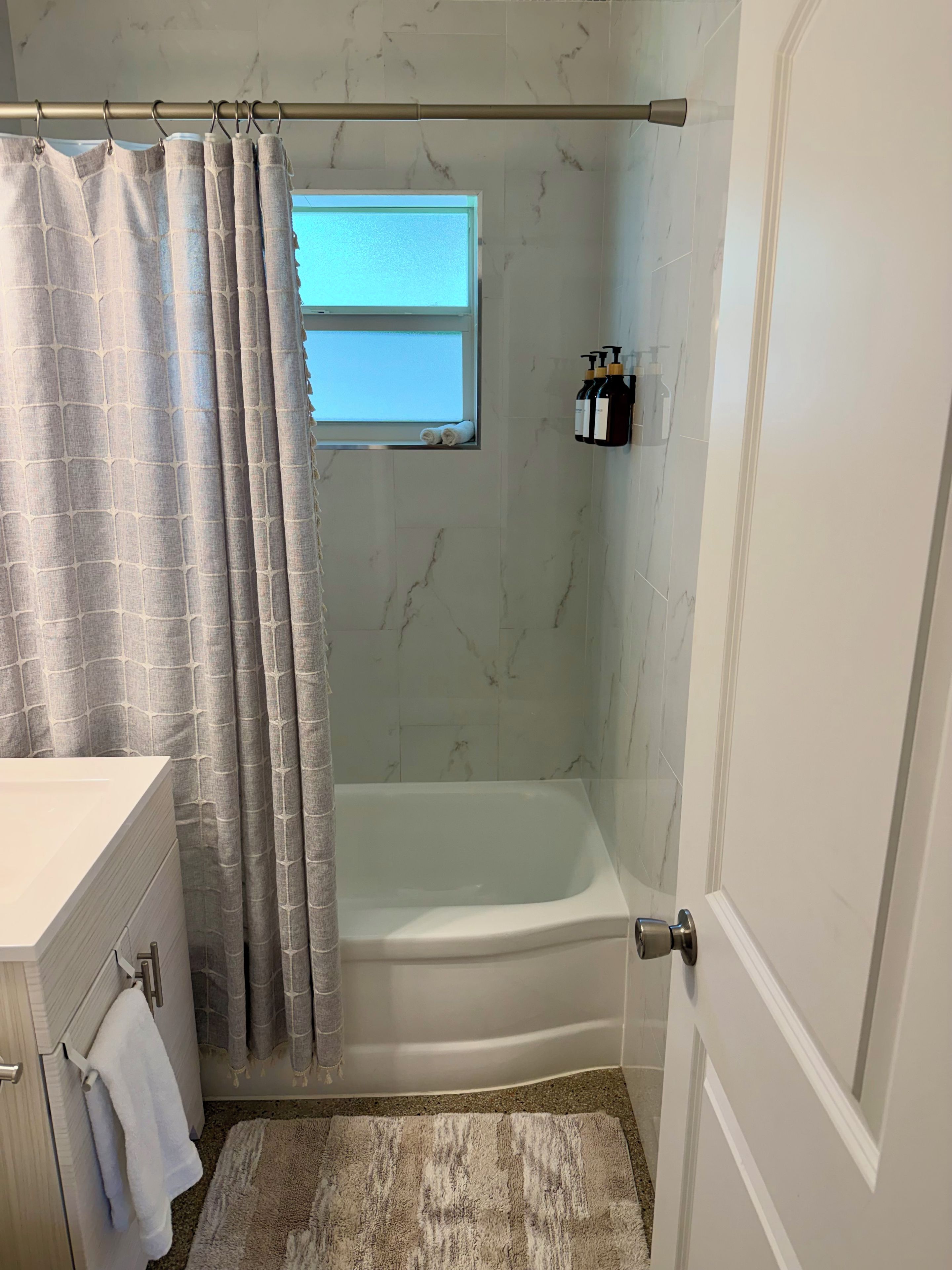 A bathroom with a bathtub, a shower curtain, a window, a towel rack, and a bottle of shampoo.