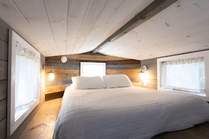 A bedroom with a large bed, wooden walls, and two wall-mounted lights. - DC57: Shell Yeah | Tiny Home | Centrally Located on Back Road (Ocracoke)