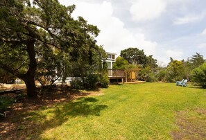 A residential property with a well-maintained lawn, a wooden deck, and a large tree providing shade. - DC57: Shell Yeah | Tiny Home | Centrally Located on Back Road (Ocracoke)