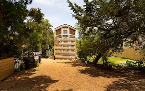 A wooden playhouse with a sloped roof and a window, surrounded by trees and a gravel path. - DC57: Shell Yeah | Tiny Home | Centrally Located on Back Road (Ocracoke)