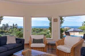 A room with a sofa, two wicker chairs, a glass coffee table, and large windows offering a view of the sea and greenery. - Casa Carpe Diem - 5 Bed Villa With Panoramic Sea Views & Private Pool (Carvoeiro)