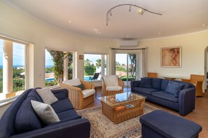 Un salon spacieux avec un grand canapé, un canapé plus petit et une table basse. On y trouve des fauteuils en osier et une table avec un plateau en verre. La pièce est dotée de grandes fenêtres offrant une vue sur l’extérieur.