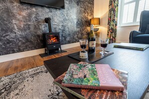 A cozy room with a wood-burning stove, a table with a book and glasses of wine, and a window with curtains. - Luxury Loft Above The Chipping Steps, Tetbury  (Tetbury)