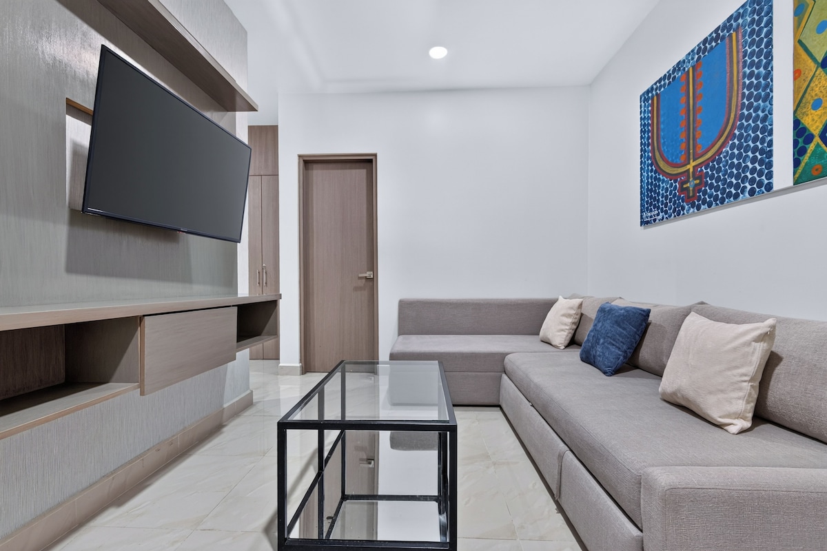 Uma sala de estar moderna com televisão de tela plana, uma mesa de centro de vidro, um sofá cinza com almofadas e uma porta de madeira.