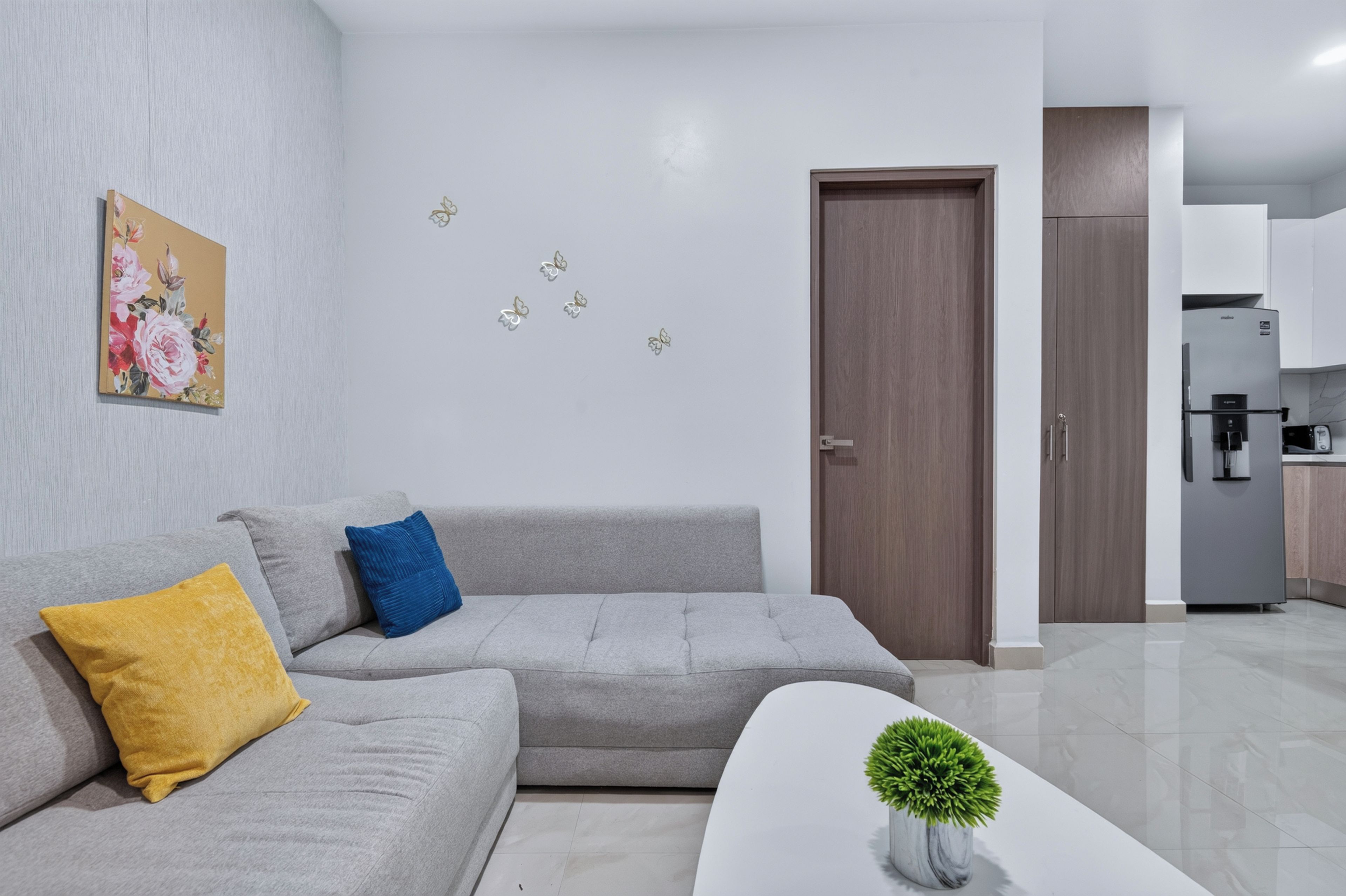Una sala de estar moderna con un sofá gris, una almohada amarilla, una almohada azul, una mesa de centro blanca con una planta, y una cocina al fondo.