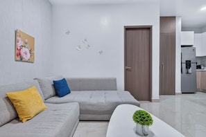 Uma sala de estar moderna com um sofá cinza, um travesseiro amarelo, um travesseiro azul, uma mesa de centro branca com uma planta, e uma cozinha ao fundo.