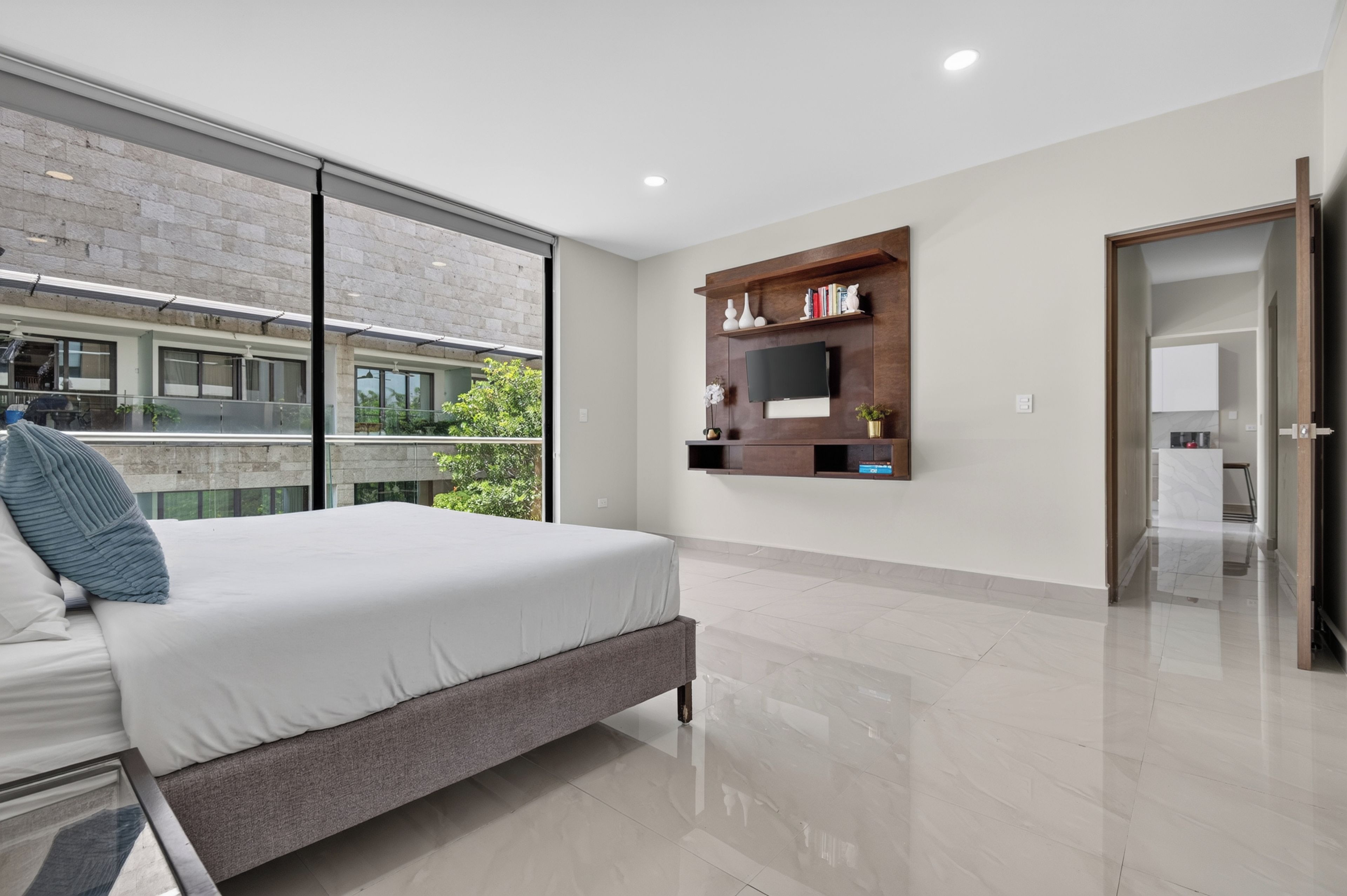Un dormitorio moderno con una cama grande, un televisor montado y vistas de un edificio a través de un ventanal.