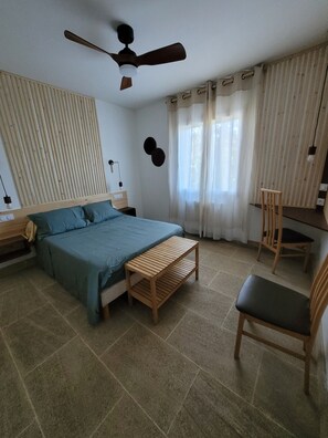 Ein Schlafzimmer mit einem Bett, einer hölzernen Sitzbank, einem Stuhl, einem Deckenventilator und einem Fenster mit Vorhängen.