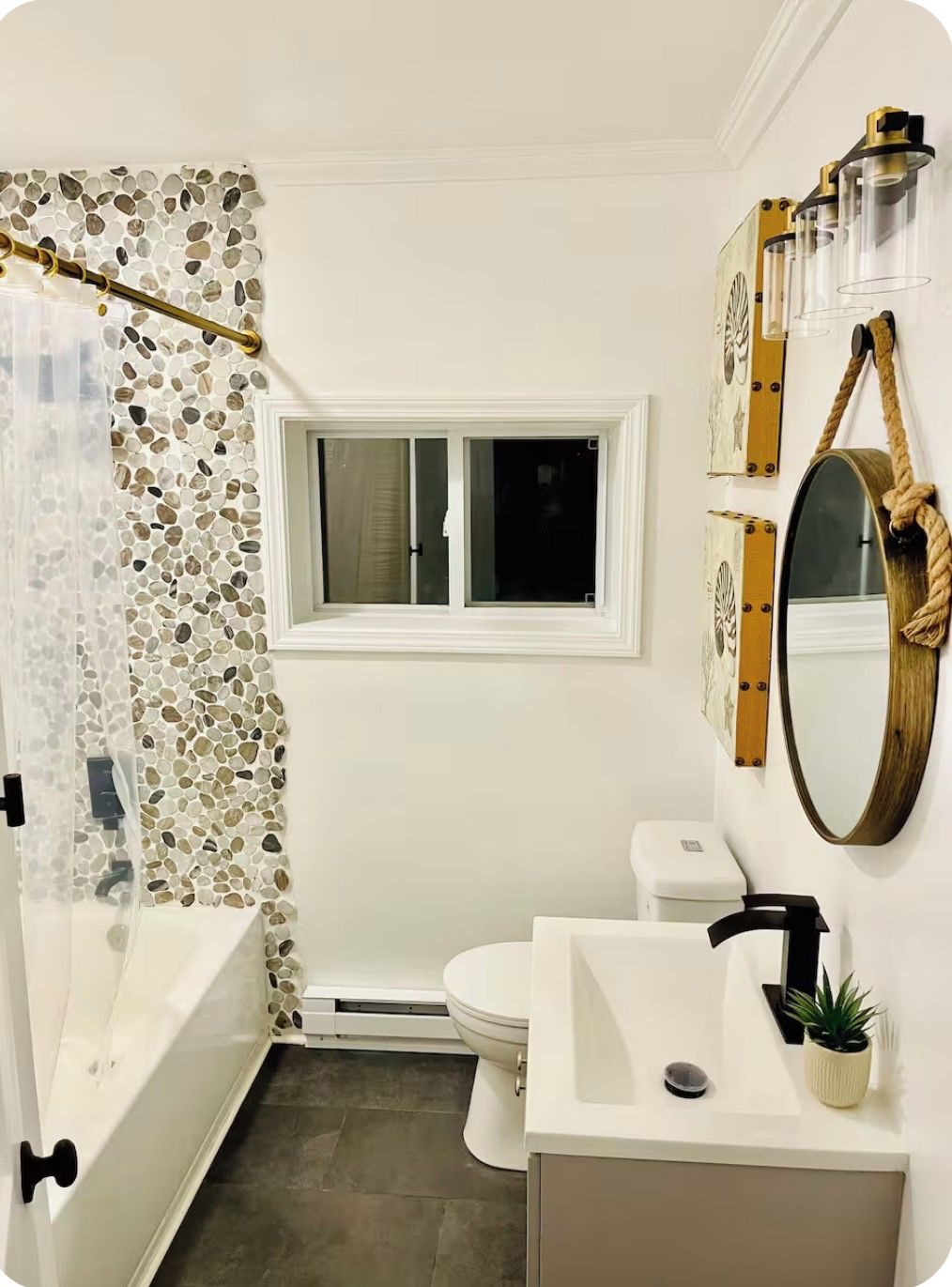 Une salle de bain avec une baignoire, des toilettes et un lavabo. Il y a une fenêtre au-dessus des toilettes et un rideau de douche à la texture granuleuse.