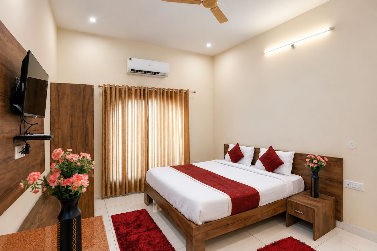 Habitación de hotel con cama, televisor montado en la pared, equipo de aire acondicionado, cabecera de madera, mesita de noche y un jarrón con flores.
