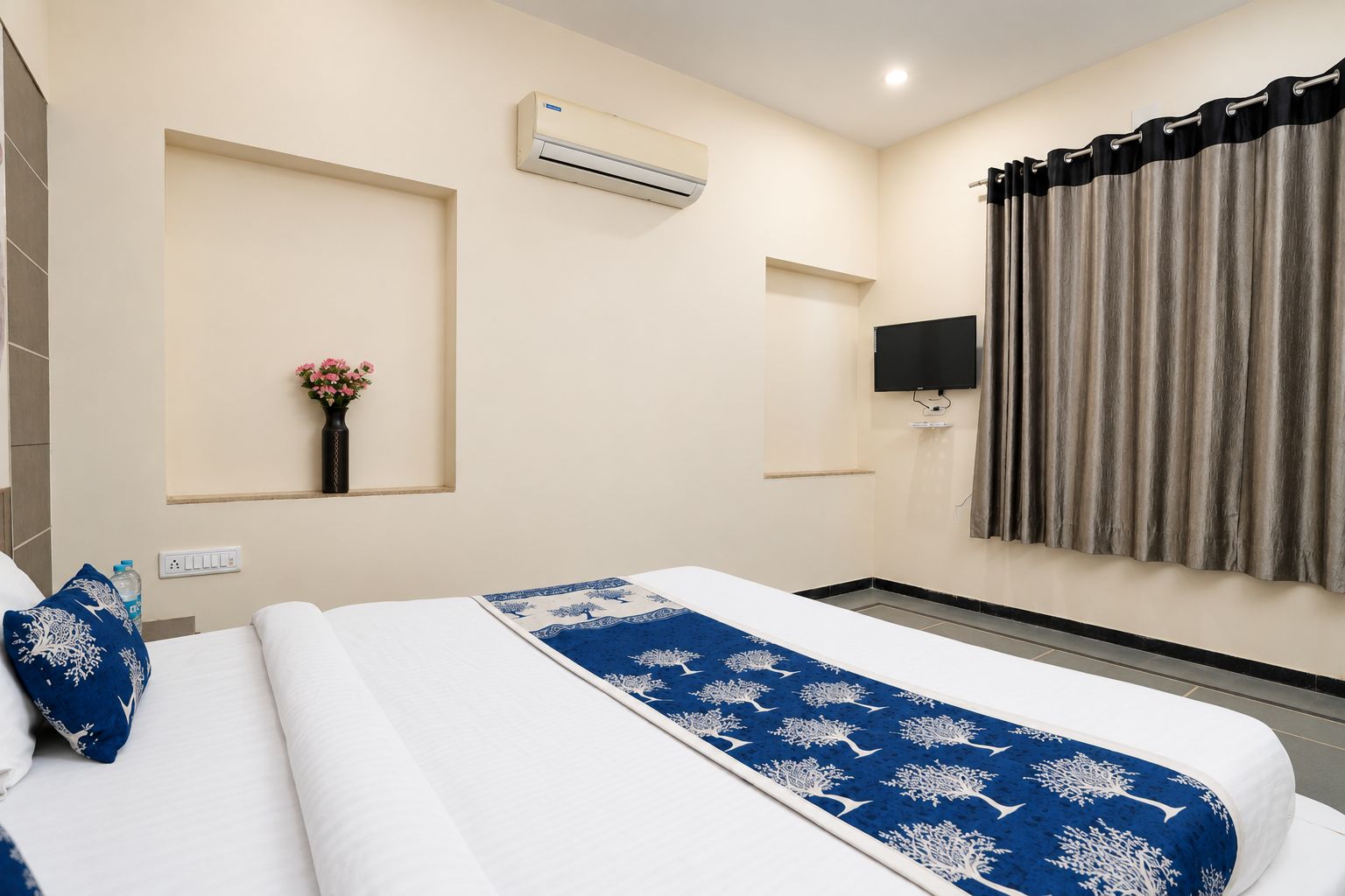 Habitación de hotel con una cama, un televisor de pantalla plana, un equipo de aire acondicionado, una ventana con cortinas y un jarrón con flores.
