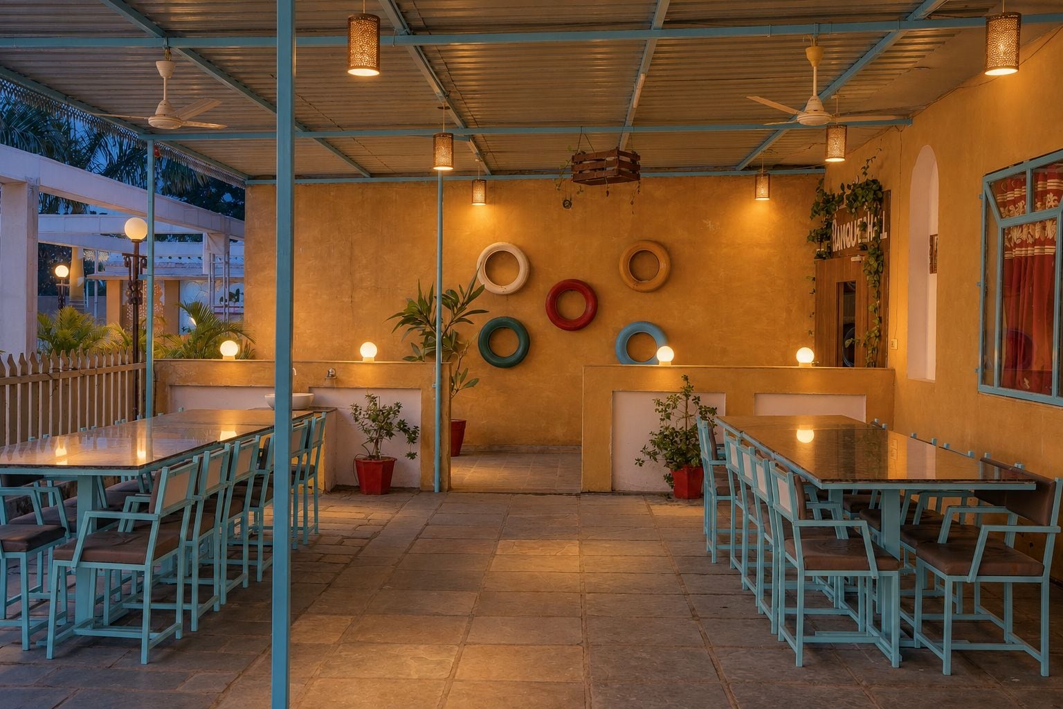 Un área de comedor al aire libre con mesas y sillas, una pared decorada con neumáticos colgantes y plantas en macetas.