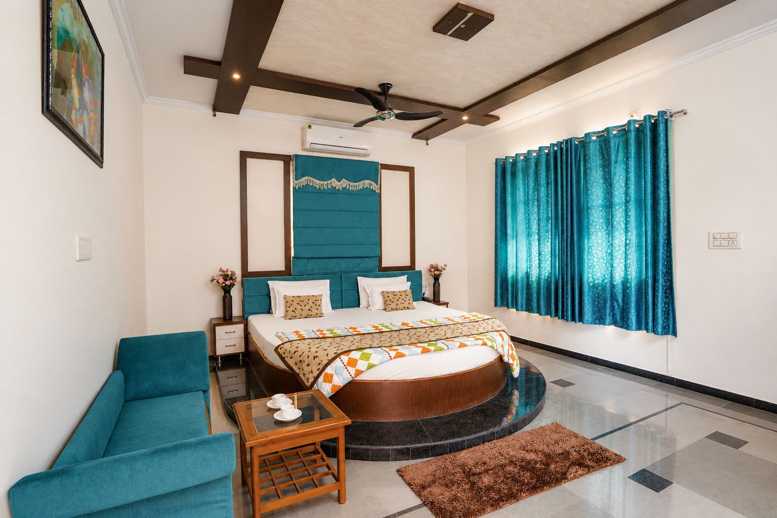 Una habitación de hotel con cama, zona de estar, una mesita pequeña, ventilador de techo y ventana con cortinas.