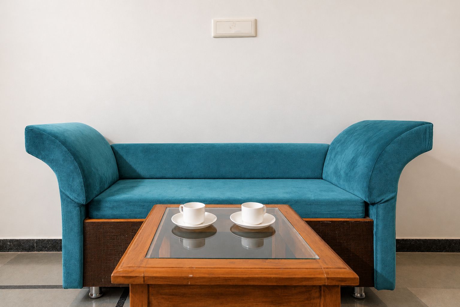Un sofá azul verdoso con una mesa de centro de vidrio, dos tazas blancas sobre la mesa, y un interruptor de luz en la pared.