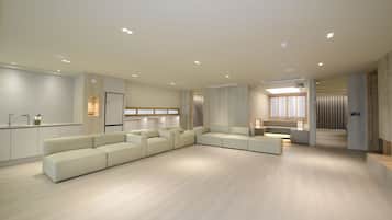 Una sala moderna con un amplio sofá modular en forma de L, una cocina integrada y una zona de estar con una mesa pequeña y sillas.