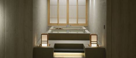 A minimalist bathroom with a built-in sink, a window with vertical blinds, and a small step leading to a pebble-lined area.