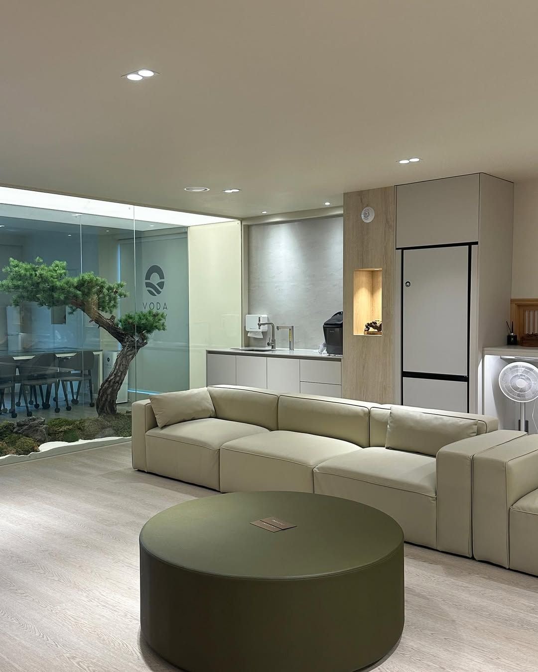 A modern living room with a sectional sofa, a round ottoman, a glass wall with a bonsai tree, a kitchen area with a refrigerator, and recessed ceiling lighting.