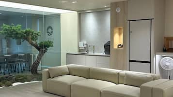 A modern living room with a sectional sofa, a round ottoman, a glass wall with a bonsai tree, a kitchen area with a refrigerator, and recessed ceiling lighting.