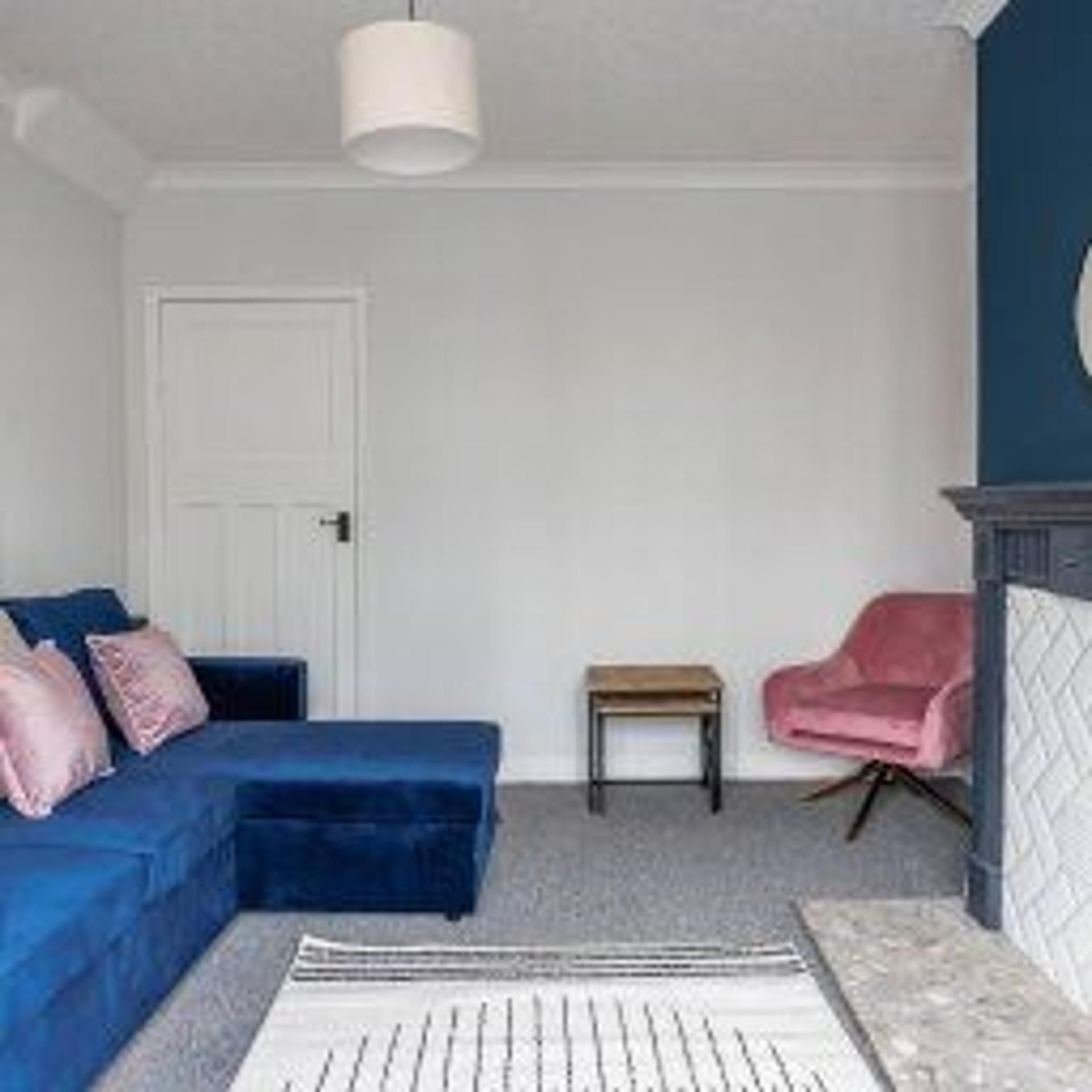 Quarto com sofá azul, mesa pequena, cadeira rosa e porta branca.