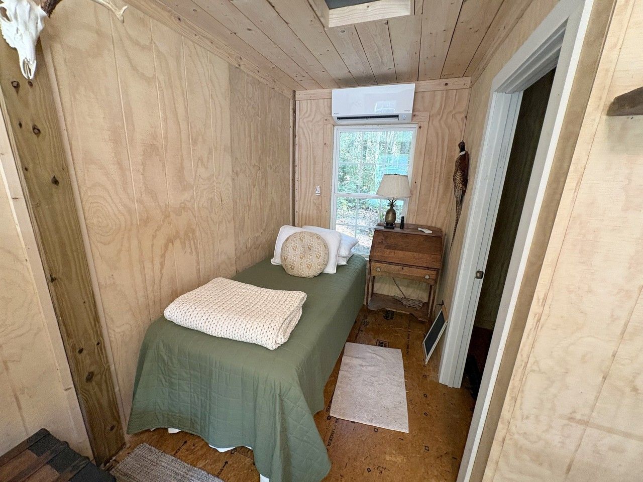 Une petite chambre à coucher, chaleureuse et accueillante, avec un lit simple, une table de chevet, une lampe et une fenêtre laissant entrer la lumière naturelle.
