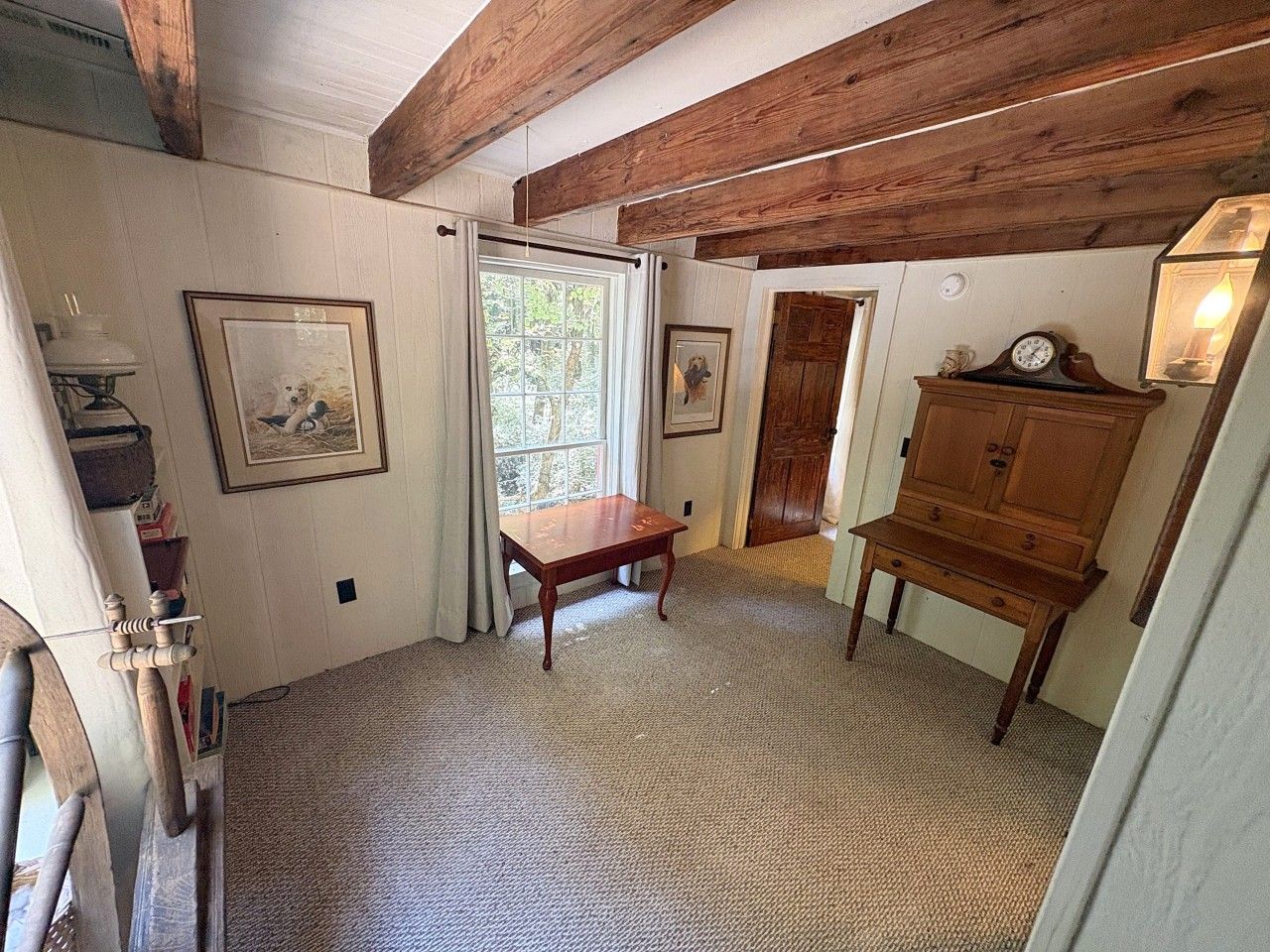 Una habitación con un banco de madera, una mesita y un armario. Hay una puerta que da a otra habitación y una ventana con cortinas.