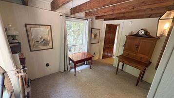 Una habitación con un banco de madera, una mesita y un armario. Hay una puerta que da a otra habitación y una ventana con cortinas.