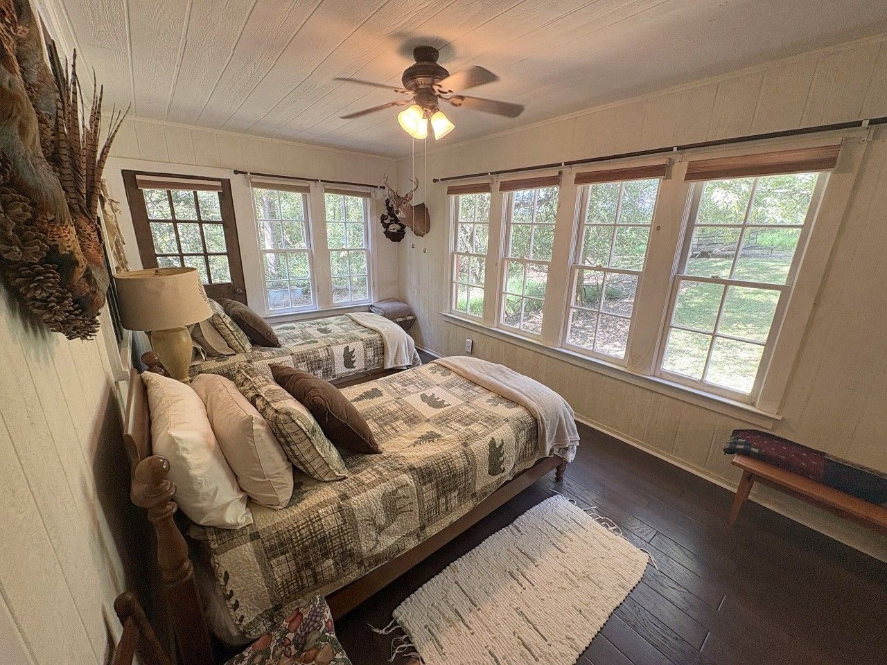 A bedroom with two beds, a ceiling fan, large windows, and a wooden floor.