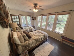 A bedroom with two beds, a ceiling fan, large windows, and a wooden floor.