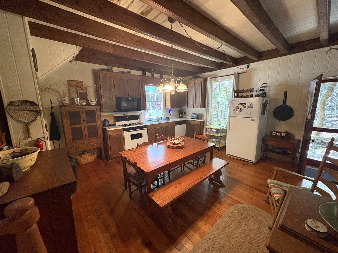Une table à manger en bois avec des bancs, un coin cuisine doté de meubles en bois, un réfrigérateur et un luminaire suspendu.