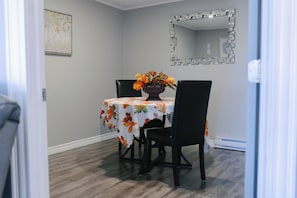 Un comedor con una mesa cubierta por un mantel estampado con motivos de hojas, dos sillas, un florero con flores y un espejo en la pared.