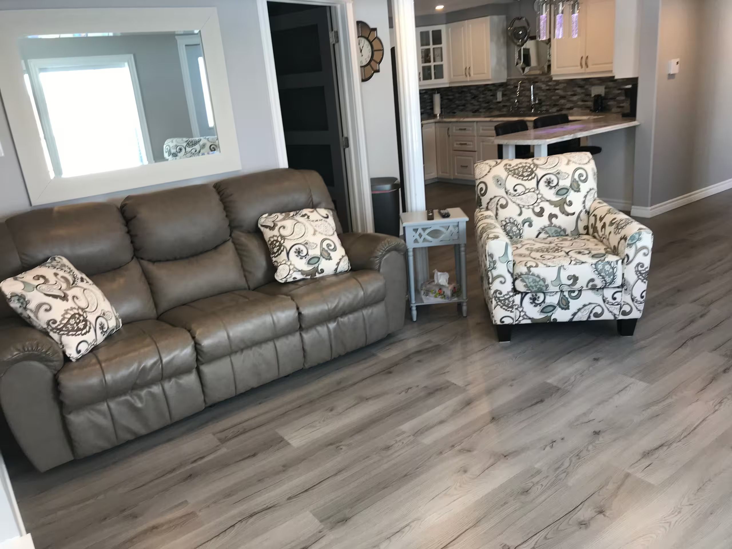A living room with a brown leather sofa and a patterned armchair, a small side table, and a view into the kitchen area.