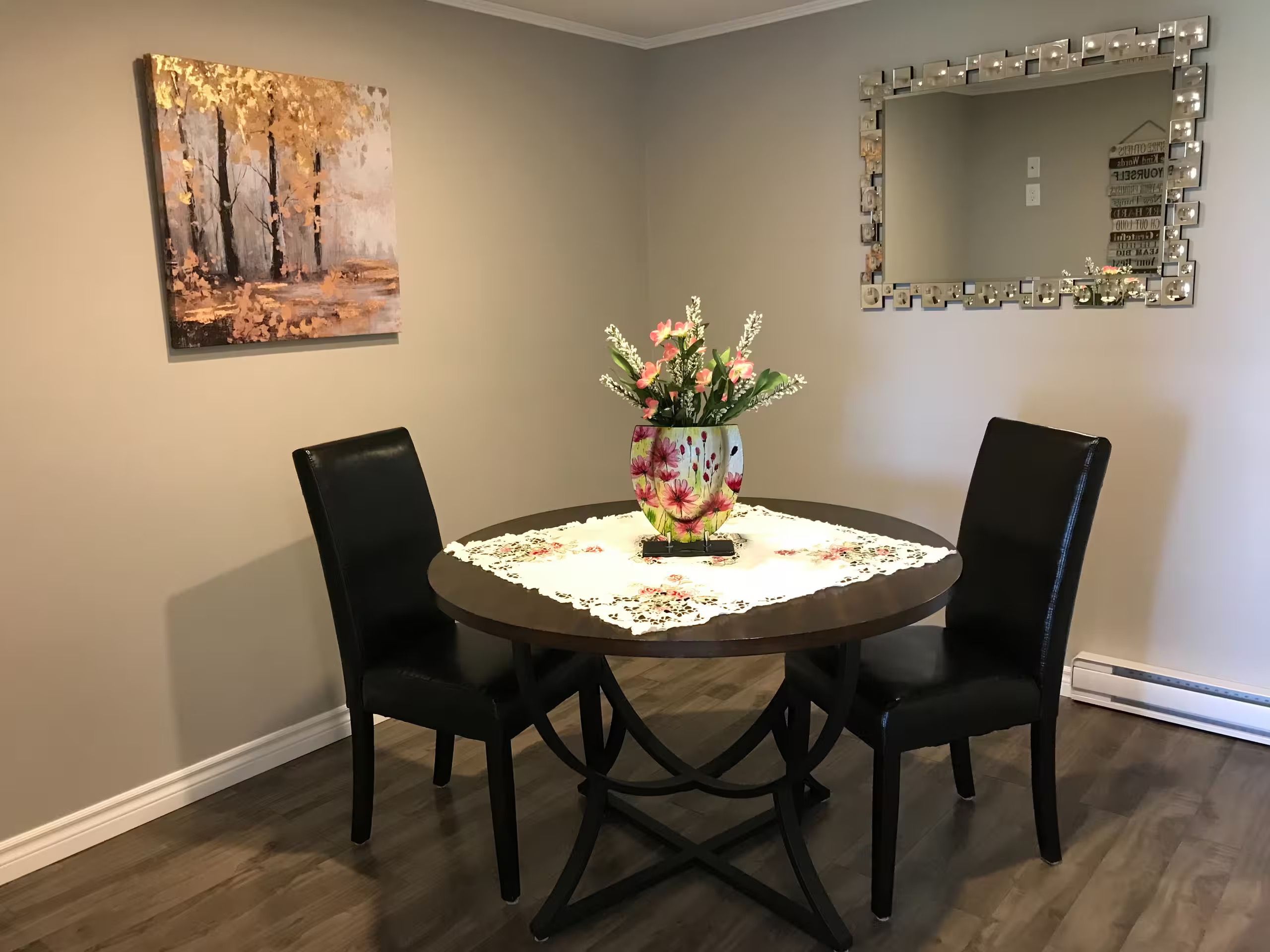 A dining area with a round table, two chairs, a floral vase, a painting, and a mirror on the wall.