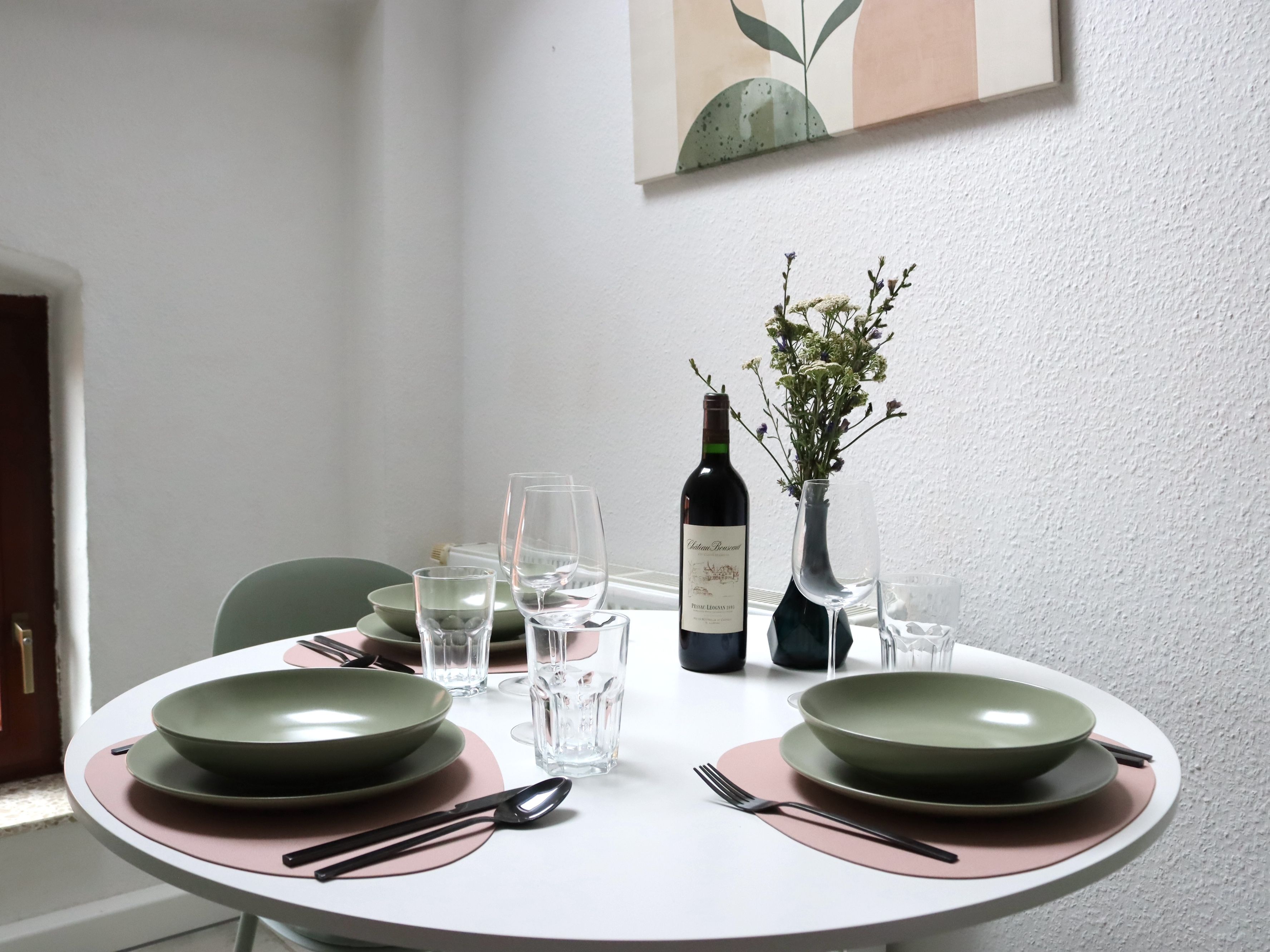 A dining table set with green plates, clear glasses, and cutlery, a bottle of wine, and a vase with flowers.