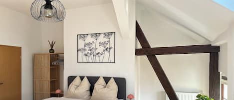 Une chambre à coucher comprenant un lit, une table de chevet, une lampe, un fauteuil vert, une armoire en bois et une œuvre d’art murale.