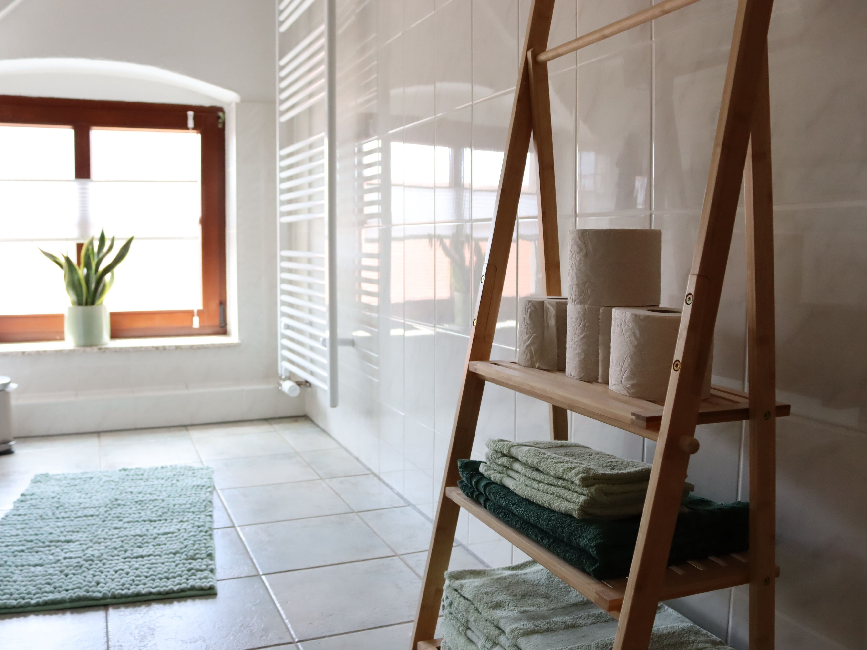 Una habitación con una estantería de madera con escalera que sostiene toallas, un tapete verde y una planta en maceta cerca de una ventana con persianas.
