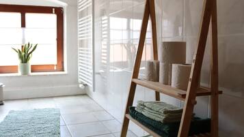 Una habitación con una estantería de madera con escalera que sostiene toallas, un tapete verde y una planta en maceta cerca de una ventana con persianas.