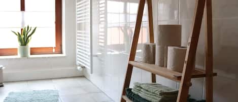 Una habitación con una estantería de madera con escalera que sostiene toallas, un tapete verde y una planta en maceta cerca de una ventana con persianas.