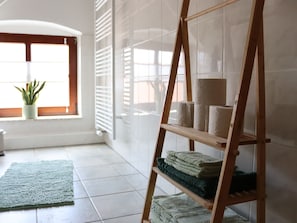 Una habitación con una estantería de madera con escalera que sostiene toallas, un tapete verde y una planta en maceta cerca de una ventana con persianas.