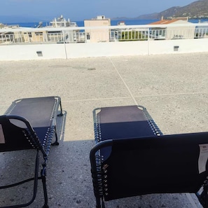 Twee lounge stoelen op een terras op het dak, met uitzicht op zee en gebouwen.