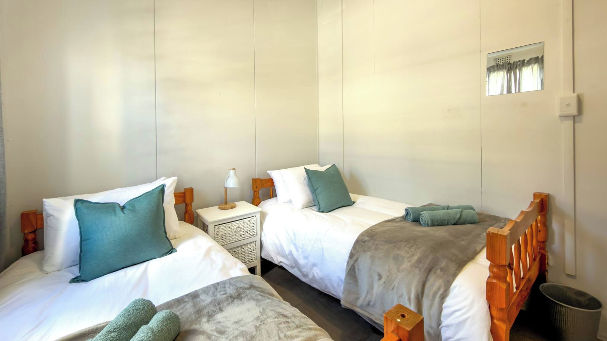 Une chambre avec deux lits, chacun équipé d’un oreiller et d’une couverture, une table de chevet avec une lampe et une petite fenêtre.