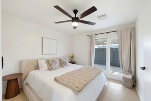 Ein Schlafzimmer mit einem Bett, einem Deckenventilator, einem Fenster mit Vorhängen und einem an der Wand hängenden Bild.