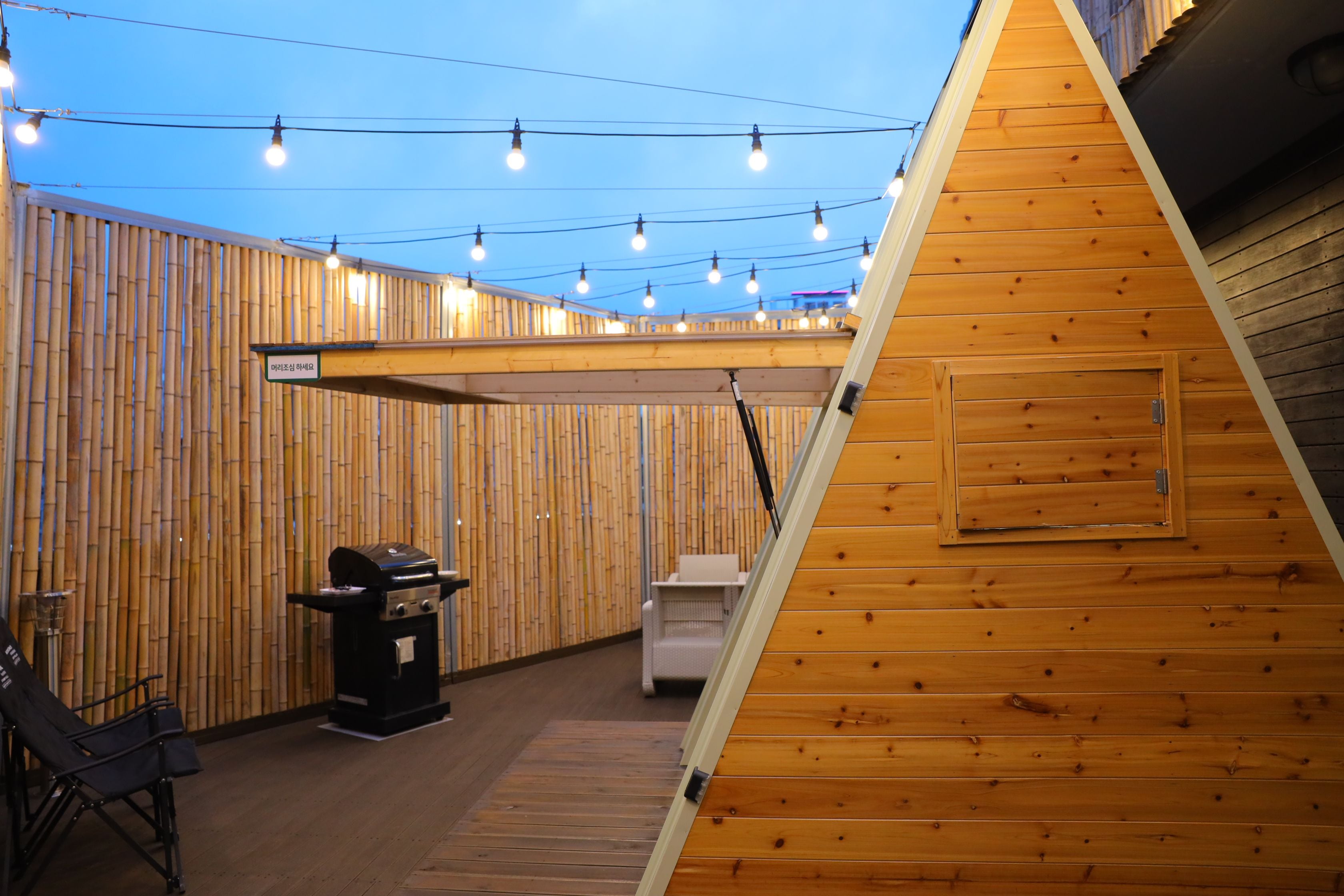 Una estructura de madera con techo inclinado, una parrilla y una zona de estar con fondo de bambú.