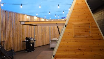 Una estructura de madera con techo inclinado, una parrilla y una zona de estar con fondo de bambú.
