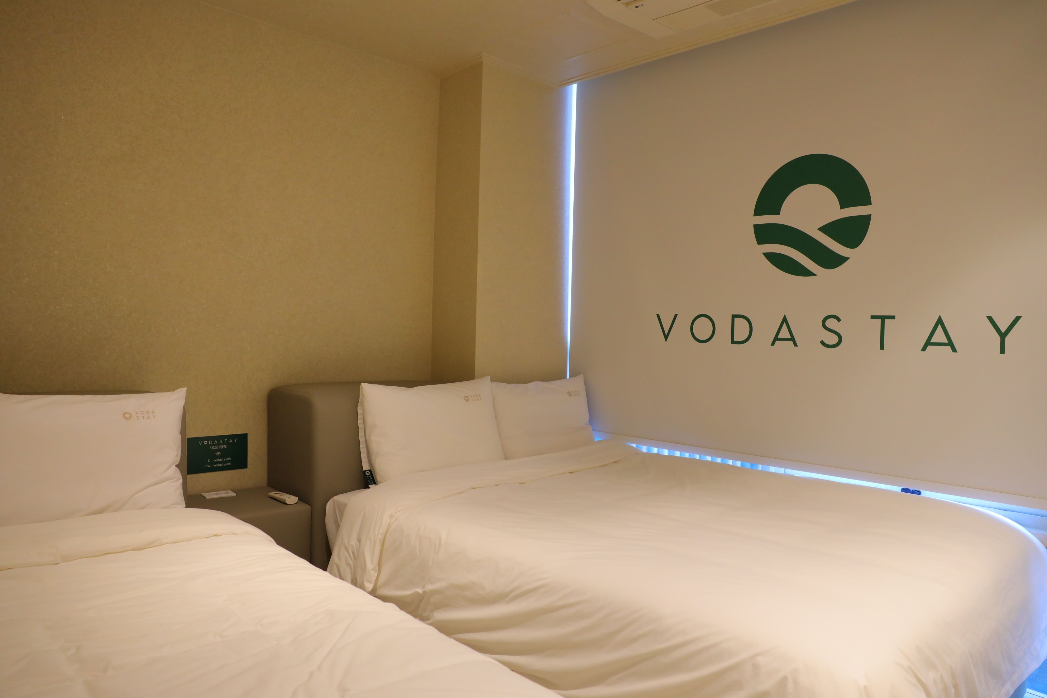 Een hotelkamer met twee bedden, een minibar en een logo van VODASTAY op de muur.