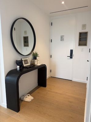 A hallway with a black console table, a round mirror, a potted plant, a framed picture, and a pair of slippers. - Prime Manhattan 2BR, 2BA apt with gym, great amenities & unbeatable location. (New York)