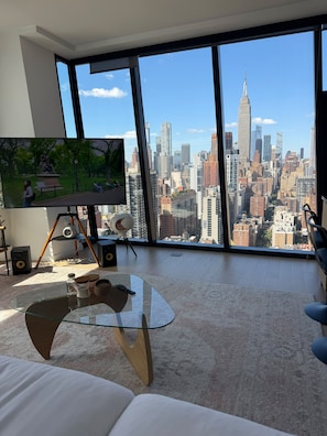 A modern living room with a large TV, a glass coffee table, and a view of a city skyline. - Prime Manhattan 2BR, 2BA apt with gym, great amenities & unbeatable location. (New York)